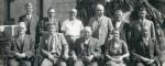 Two rows of people, three standing behind three sitting. In the middle of the front row is a young woman in a dress with short hair, the rest of the people or older men wearing suits.